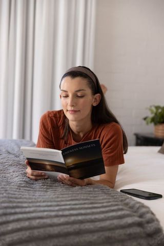 Woman lying on stomach reading endless sunsets book in cozy sunlit bedroom with smartphone