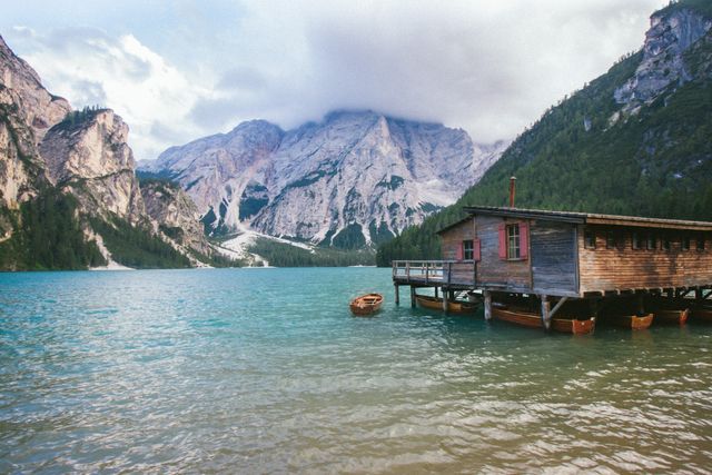 Beautiful rustic boathouse on calm, clear lake surrounded by majestic mountains and lush forests. Perfect for travel brochures, nature-themed articles, outdoor adventure promotions, and wellness relaxation visuals.