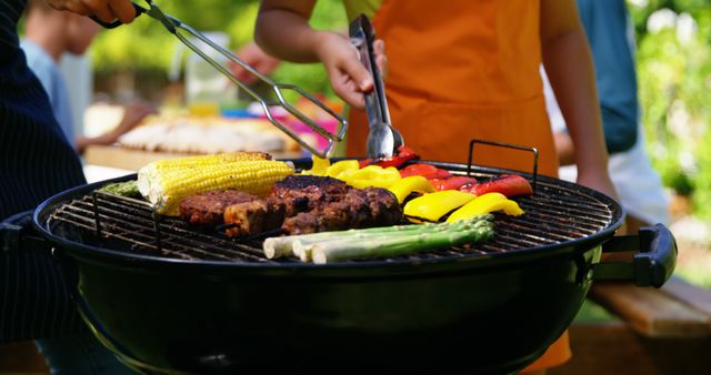 People grilling vegetables and meat on a barbecue outside in a park or backyard. Various vegetables such as corn, asparagus, and bell peppers are on the grill alongside grilled meat. Ideal for use in advertisements, cookout event promotions, and articles related to summer activities, healthy eating, and outdoor gatherings.