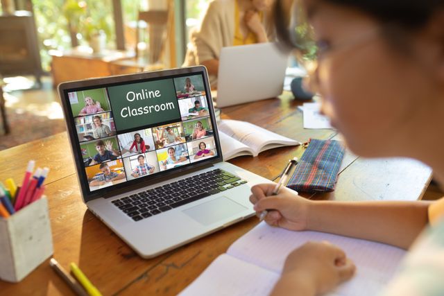 Asian teenage girl engaged in an online class with a laptop at home, writing notes in a notebook. Ideal for illustrating topics related to online education, virtual classrooms, distance learning, and the use of technology in modern education systems.