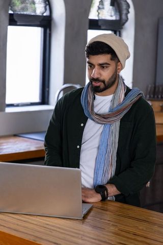 Young man wearing beanie and scarf working on laptop at cozy cafe counter