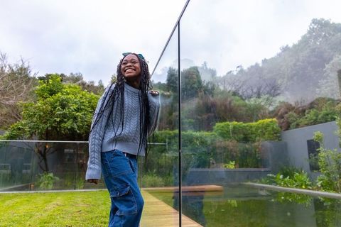 African american teen girl leaning on glass railing, smiling on backyard deck by pond