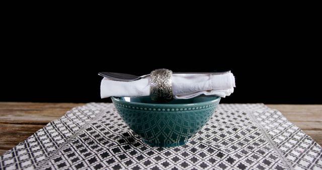 An elegant table setting featuring a silver napkin ring, a neatly folded white napkin, and a green ceramic bowl. The items are placed on a geometric patterned placemat on a wooden table. This image is perfect for use in dining decor promotion, table setting arrangements, and stylish dinnerware advertisements. Ideal for websites, blogs, menus, and social media posts that focus on fine dining and home decor.