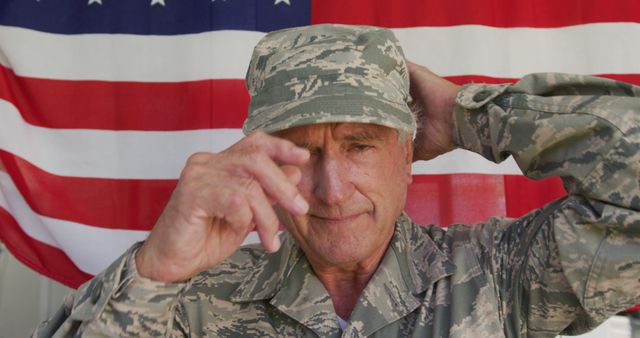 This image depicts a senior military veteran dressed in a camouflage uniform, adjusting his cap while standing in front of a large American flag. His expression is solemn and respectful, emphasizing themes of honor, service, and patriotism. This image can be used in advertisements, articles, or educational material related to military service, veteran affairs, patriotic events, national holidays like Memorial Day and Veterans Day, or topics of national pride.