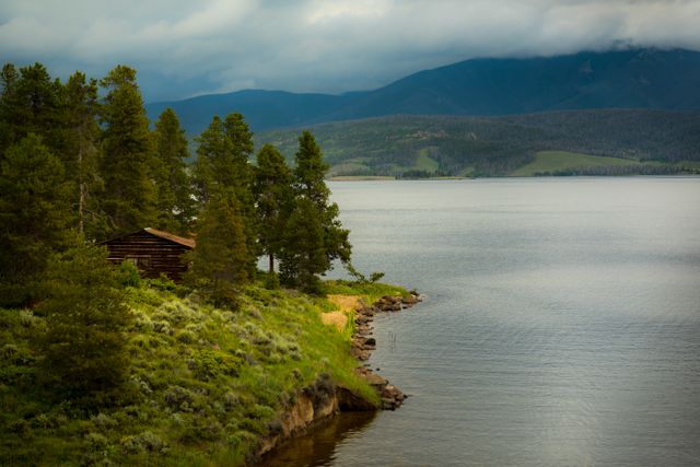 Peaceful lake setting with a quaint cabin nestled among lush pine trees and mountainous backdrop provides a sense of tranquility and connection with nature. Ideal for use in travel advertising, nature retreat brochures, environmental conservation campaigns, or outdoor adventure promotions.