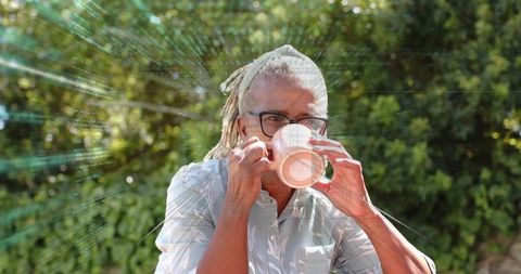 Mature woman with dreadlocks sipping coffee in sunlit garden with cracked glass overlay