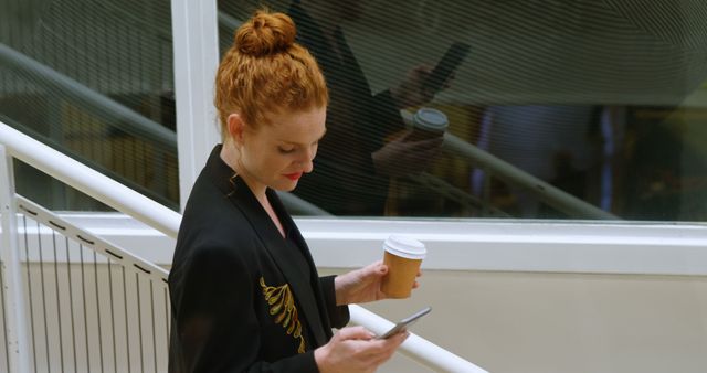 Young Caucasian woman checks her phone in an office setting. She balances work with a coffee break, staying connected on the go.