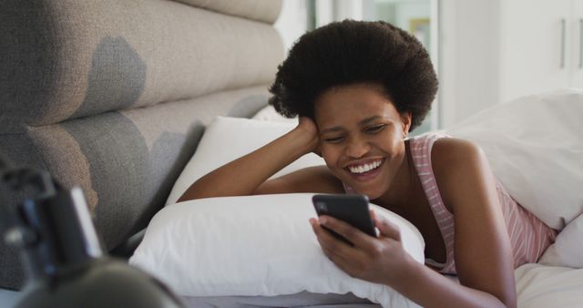Woman lying on bed comfortably, laughing while holding smartphone. Picture can be used for themes like relaxation, modern technology, leisure, or home life. Ideal for marketing campaigns, website banners, blogs on technology or home living, and social media posts about communication and happiness.