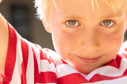 Smiling blonde boy raising arm wearing red white striped shirt sunlit closeup portrait