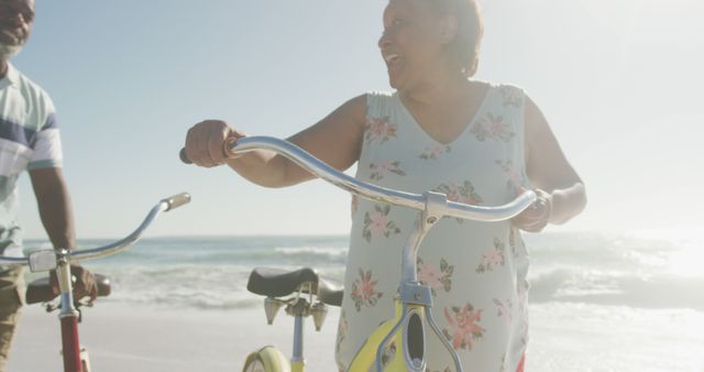 Smiling senior african american couple walking with bicycles on sunny beach. healthy, active retirement beach holiday.