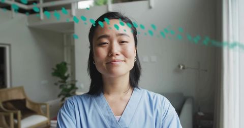 Confident Korean nurse standing in modern living room wearing light-blue scrubs smiling