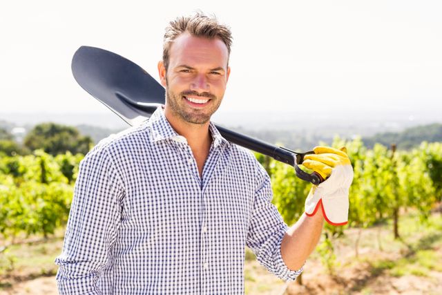 Young man standing in vineyard holding shovel over shoulder, smiling. Wearing plaid shirt and gloves. Ideal for use in agricultural promotions, farming advertisements, rural lifestyle content, and gardening blogs.