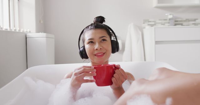 Image of smiling biracial woman with headphones and red cup of tea in bathtub in bubble bath. Health and beauty, leisure time, domestic life and lifestyle concept.