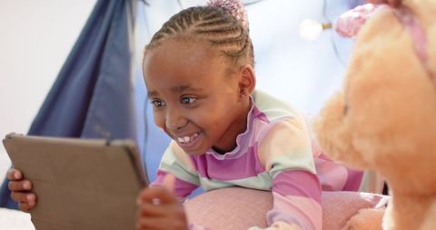 Smiling african american girl playing with tablet in pastel teepee bedroom with teddy bear