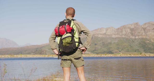 Male hiker with backpack standing by mountain lake, enjoying scenic view. Ideal for use in travel brochures, adventure blogs, camping gear advertisements, outdoor activity pamphlets, and nature exploration articles. Highlights the concept of adventure, exploration, and solitude in natural settings.