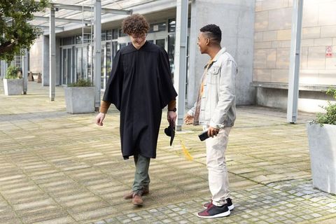 Young male graduate walking with friend on campus courtyard holding cap and phone