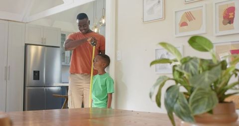 Dad measuring son height in modern kitchen marking growth milestone with tape measure