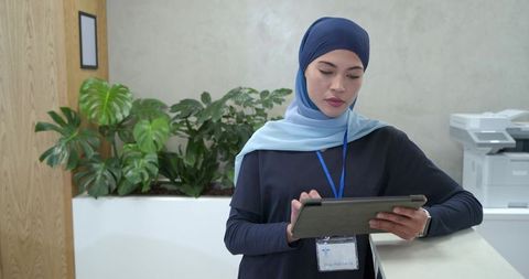 Receptionist in hijab holding tablet and showing id badge at modern office reception desk