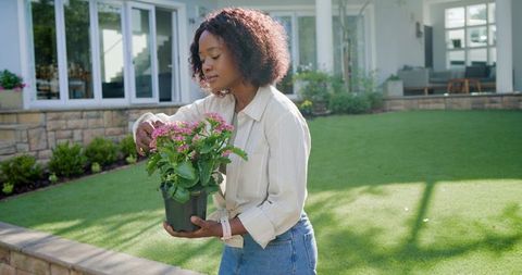 Woman tending potted pink flowers on sunny backyard lawn, casual at-home gardening