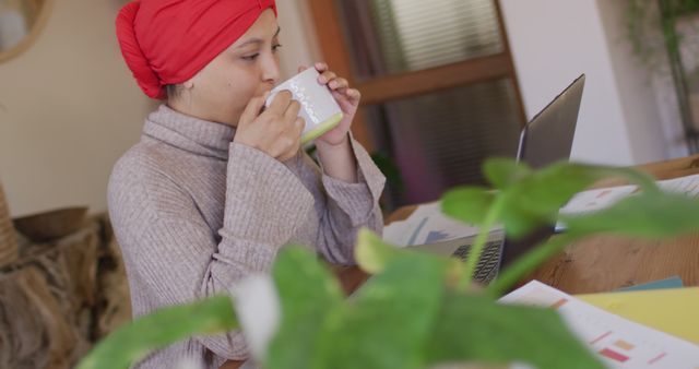 Image of biracial woman in hijab sitting at desk at home working on laptop, drinking coffee. Remote working, communication, inclusivity and domestic life.
