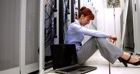 Sitting IT technician troubleshooting laptop between server racks in data center