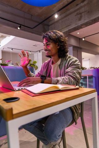 Asian man smiling and gesturing while working on laptop in modern coworking space