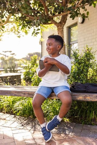 Smiling african american boy holding tablet on bench in sunny courtyard by school