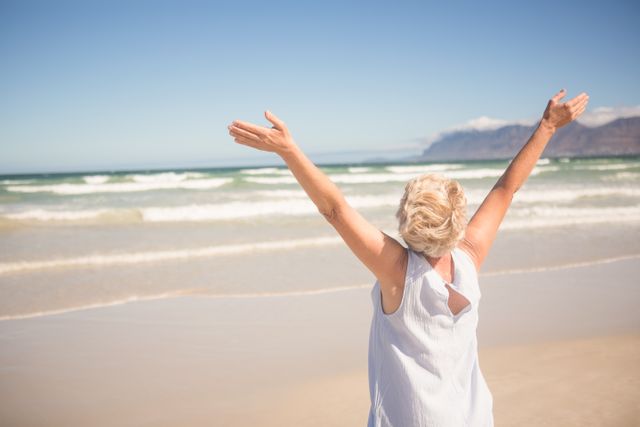 This image captures a woman standing on the shore with her arms raised, embracing the freedom and joy of being at the beach. Ideal for use in travel brochures, wellness blogs, advertisements promoting beach destinations, or any content related to relaxation and happiness.
