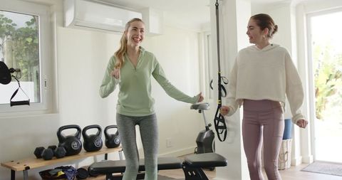 Female friends exercising together in sunlit home gym with trx, kettlebells, weight bench