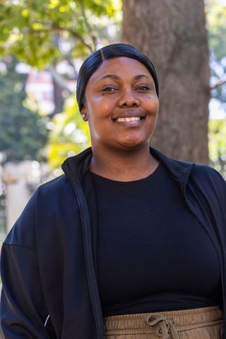 African woman smiling and standing in sunlit park wearing black jacket and casual pants