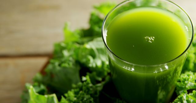 A glass of vibrant green juice is surrounded by fresh leafy greens on a wooden surface, suggesting a healthy, nutrient-rich drink. The focus on the juice and greens emphasizes the importance of fresh produce in a balanced diet.