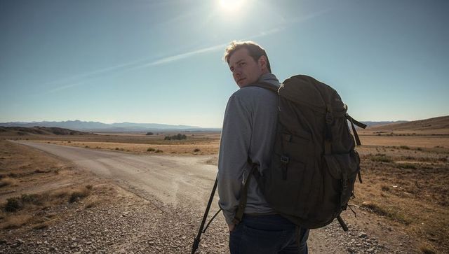Hiker looking back at crossroads in sunlit grassland carrying large backpack and trekking pole
