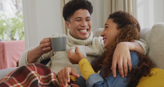 Image of happy biracial couple siting on sofa under blanket and drinking coffee. Domestic lifestyle and leisure time at home.
