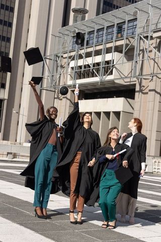 Diverse female graduates tossing mortarboards and holding diplomas in city plaza