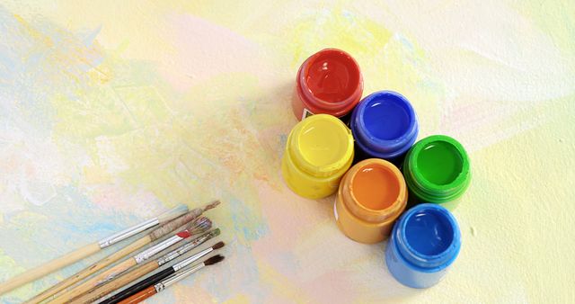 Open jars of colorful paint are arranged on a pastel background smeared with various hues, with copy space. Paintbrushes lie nearby, suggesting an artistic process or a painting session in progress.