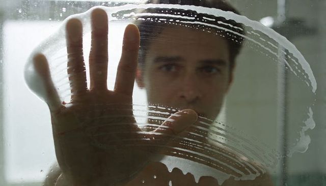 Man pressing hand on soapy shower glass, silhouette standing behind steamed partition