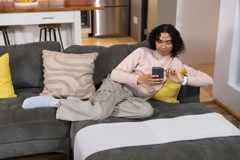Non-binary adult relaxing on gray sectional scrolling smartphone in cozy living room