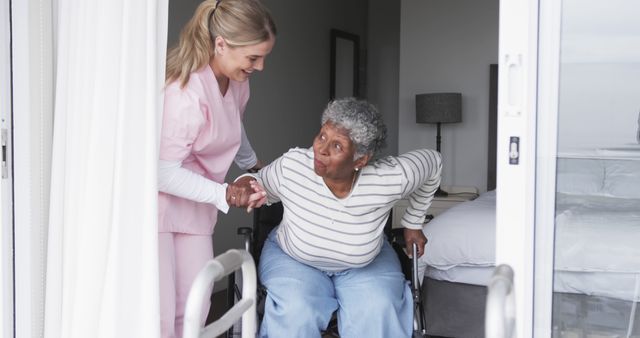 Senior african american woman in a wheelchair receiving assistance from a healthcare worker inside a nursing home. The healthcare worker, dressed in pink scrubs, is helping the woman with her arm as she prepares to stand or transfer. This image can be used to represent elderly care, rehabilitation, nursing homes, patient care, and the compassionate relationship between healthcare providers and their patients.