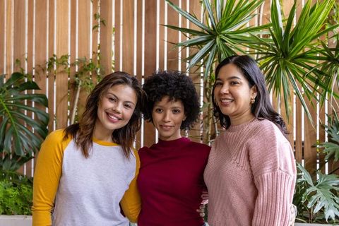 Multicultural women smiling and embracing on plant-filled patio with wooden slat backdrop