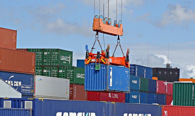 Crane lifting a blue shipping container onto stacked cargo boxes at busy port. Ideal for illustrating global logistics, international trade, commercial transportation, and supply chain management. Useful in articles about shipping industry, port activities, or logistics solutions.