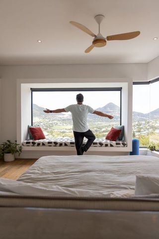 Mature man balancing on one leg on window bench overlooking valley in sunlit minimalist bedroom