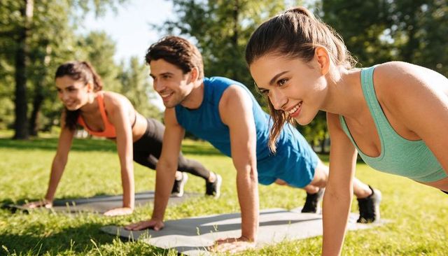 Group exercising outdoors doing plank on mats in sunny park with smiling young adults
