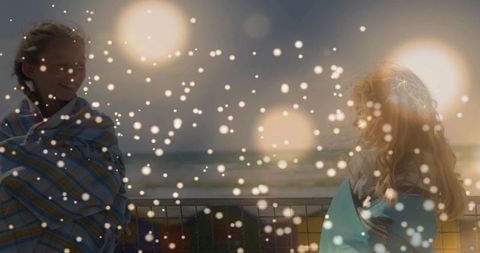 Two girls wrapped in striped teal blankets at pier railing during sunset with dreamy bokeh