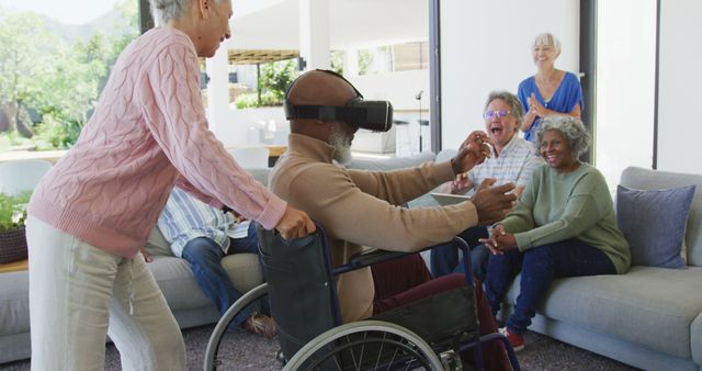 Senior man in a wheelchair enjoying virtual reality with usage of VR headset, surrounded by happy friends in a modern living room. Ideal for content related to seniors embracing technology, group entertainment, and virtual reality experiences. Useful for articles, blogs, and advertisements discussing aging populations engaging with modern advancements and inclusive tech use.