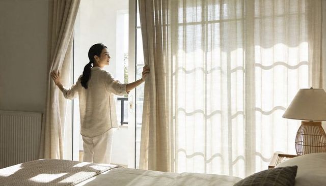 Woman pulling linen curtains at glass balcony door in sunlit minimal bedroom