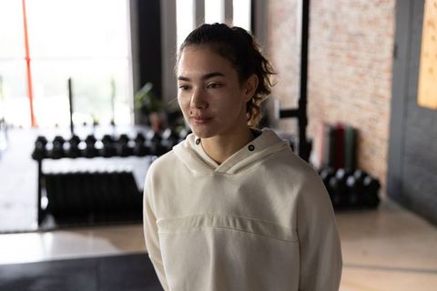 Young woman standing in gym wearing pullover hoodie near dumbbell rack and kettlebells