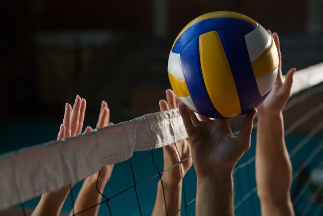 Hands of players reaching for volleyball over net in indoor court. Ideal for sports-related content, teamwork and competition themes, fitness and exercise promotions, and athletic training materials.
