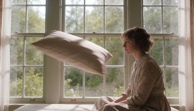Young woman sitting on sunlit window seat gazing forward while pillow floating mid-air