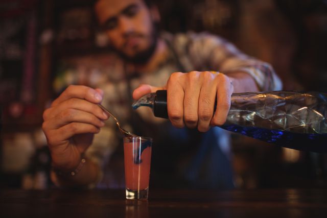 Bartender pouring a colorful cocktail into a shot glass at a bar counter. Ideal for use in articles or advertisements about nightlife, bars, mixology, and alcoholic beverages. Can be used to illustrate the art of cocktail making or to promote bar events and parties.