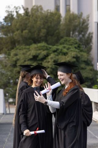 Female graduates celebrating taking selfie on campus with diplomas and mortarboards